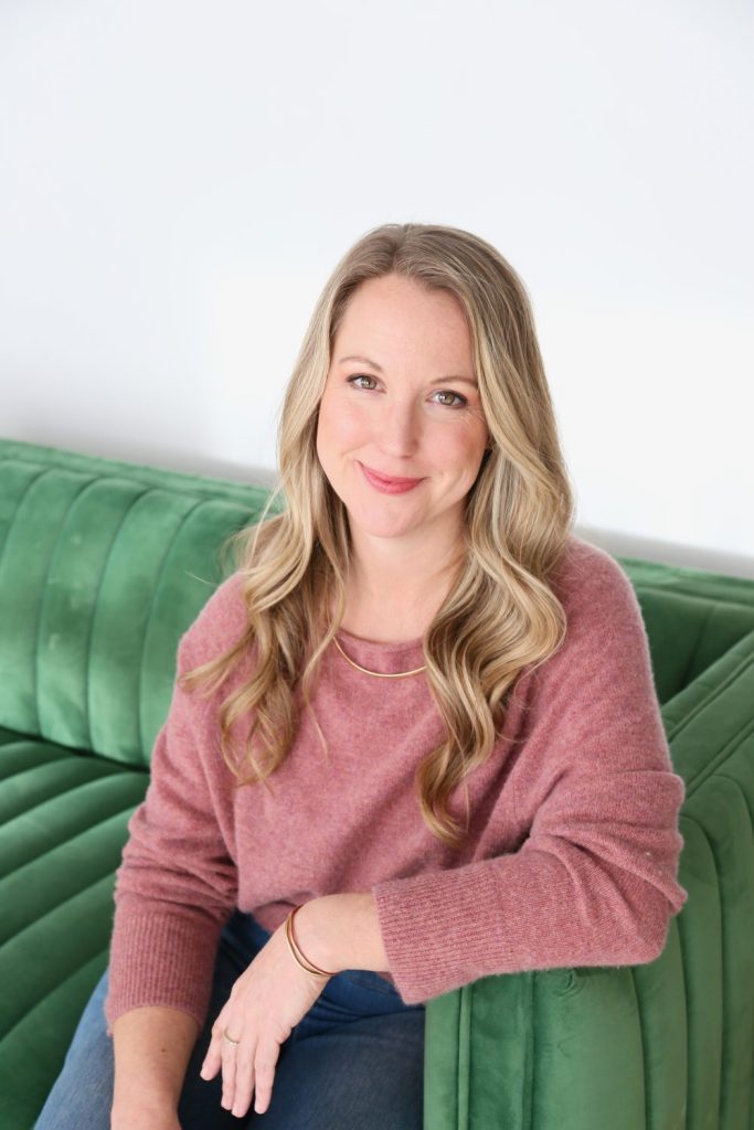 Emily Wishall smiling softly while seated on a green couch, welcoming clients into her 3-month somatic coaching program.